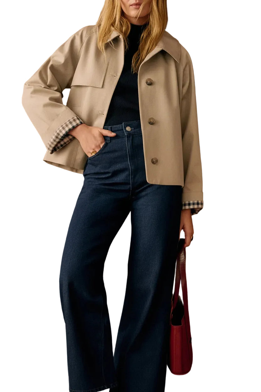 A woman models a beige cropped utility jacket with a black turtleneck and dark blue wide-leg jeans, standing against a warm white-paneled wall and holding a red handbag.
