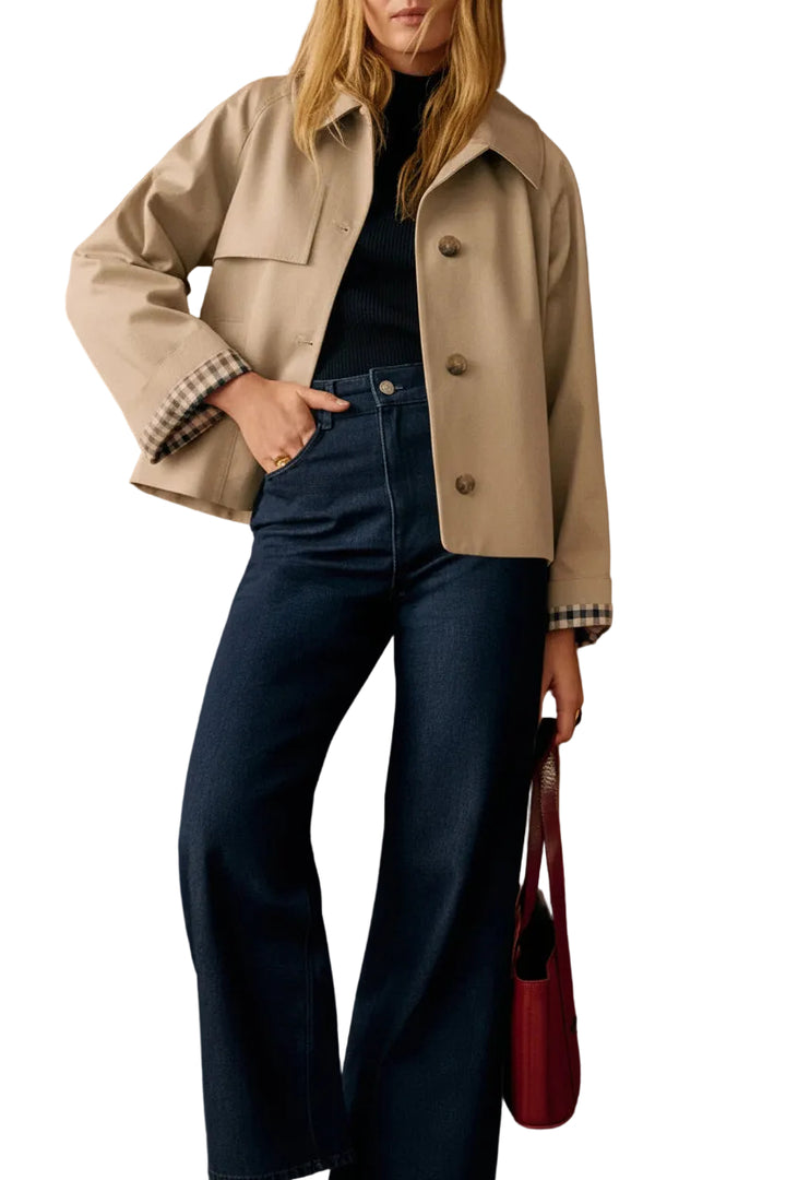 A woman models a beige cropped utility jacket with a black turtleneck and dark blue wide-leg jeans, standing against a warm white-paneled wall and holding a red handbag.