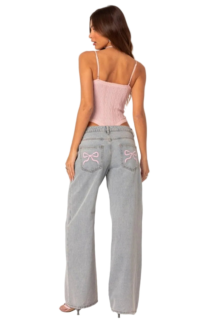 A woman shown from the back wearing a light pink ribbed camisole and light gray wide-leg jeans with pink bow details on the back pockets, against a plain light white studio background.